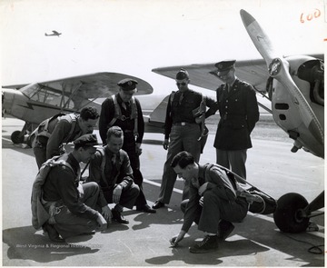 A photograph of a group of soldiers gathered around an individual drawing a diagram on the tarmac of the airstrip.