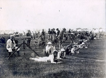 A photograph of soldiers shooting rifles, and observers with telescopes behind, at a military camp.