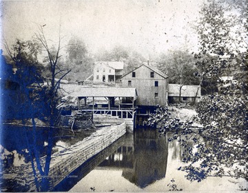 A photograph of an unidentified mill along a stream with a home in the background.