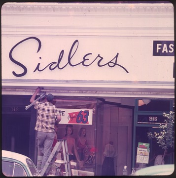 Man working on store front above display window and entrance to Sidlers Department Store in Morgantown, W. Va.