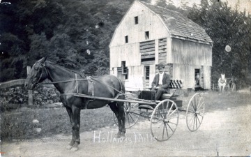 A photograph of a man sitting in a horse-drawn carriage with a mill in the background.