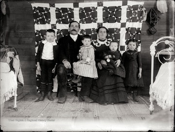 A portrait of a couple and their four children taken indoor in front of a quilt hanging on the wall.