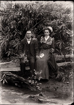 A portrait of a couple sitting on a log in front of rhododendron. 