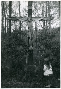 Little girl gazing up at a cross with Jesus in the woods.