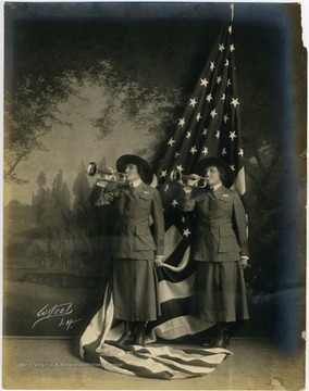Two unidentified women in military uniforms with the letters "YMCA" on the coats, stand in front of an American flag, with bugles to their lips. Judging from the style and patriotic theme, the photo was likely taken during World War I.