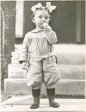 Unidentified little girl wearing hair ribbons, a romper suit and high buttoned shoes.