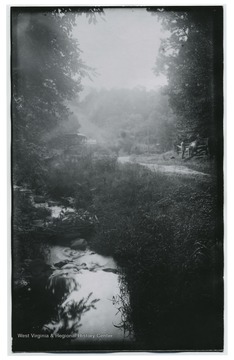 A small creek meanders through the photo.To the side is a man petting a horse. A buggy is parked by a sign that read 'Look Out for Locomotives.'  (80)D.I.C 138 appears on the back of the photo.