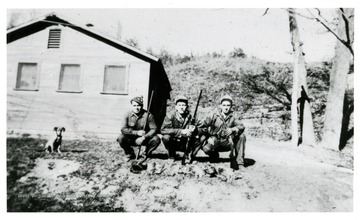 Three males squat with rifles on their shoulders; on the ground spread the hunted animals while a dog sits patiently next to the subjects.