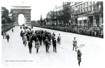 'West Virginia contingent marching on Avenue des Champs E'lysees, September 19, 1927.  Arc de Triomphe de l'Etoile in the background.  Placard bearer: Sam Stutler, Charleston.  First row, left to right: Guard of honor, Louis A. Johnson of Clarksburg, A. E. Haan of Huntington, guard of honor.  Second row includes: Dr. Gribble, C. H. Thompson, Burton Raine, Dr. Langfitt, Dr. Davis, James M. Guiher, Raine unidentified; others from Harrison County.'