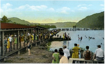 A view of Sunset Beach, people in bathing suits and in casuals relax on the pier or frolic in the water.  The Sunset Beach in Lake Lynn Pa. is located near Morgantown, W. Va. and Montchateau Hotel.