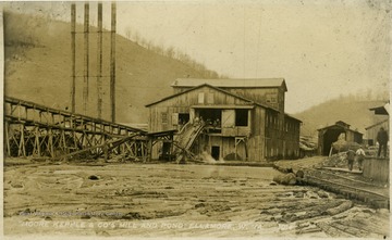 In the background, unidentified workers sit at the top of the belt which carries the logs from the pond into the mill.