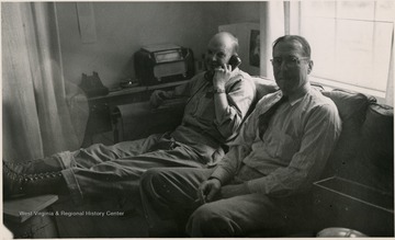 Two men posed sitting on a couch. One of the men is using the telephone.
