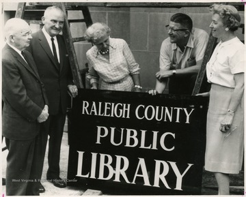 From "Beckley U.S.A." by Harlow Warren, p. 735, vol. 2. In book: "Friends of the library hang a sign: W. O. Syndor, Jr., Roy Milliron, Mrs. Nell McKenzie, Alex N. Beckinridge and Mrs. C. H. Graham, Raleigh County Librarian" (p. 735).