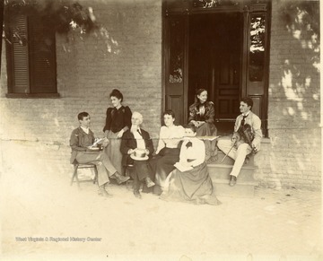 Pictured: Dr. Gregg Gibson, Sue Pendleton, Hopkins Gibson, Mr. Gregg Gibson, Robert Gibson, Lucy Pendleton, and Agnes Gibson, sitting outside on the front steps and sidewalk.  