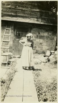Sally Miller, an elderly woman standing in front of a stone house, wearing long dress, apron and a pioneer bonnet