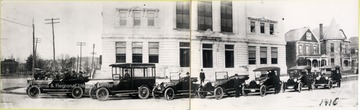 A line of automobiles, including a car of uniformed police officers, a paddy wagon and several passenger cars, parked on the street, possibly waiting to be an escort.