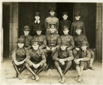 Group of young men in dress army uniforms, probably West Virginia University students, posing outside a university building. Two of the soldiers have Greek symbols printed in ink on their left shoulder.