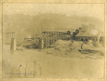Information inscribed on the back of the photograph, "Caused by 13 loaded cars breaking loose and jumping over the side of the steel span causing it to "upset"and igniting the coal dust and gas. The photograph was taken after the clean up had begun. The building to the right is the new power house and boiler room, only a small portion of the power house is visible".