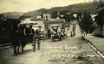 Postcard photograph of a long line of buggies and wagons in a 4th of July Industrial Parade in St. Mary's, West Virginia. See original for correspondence written on the back.