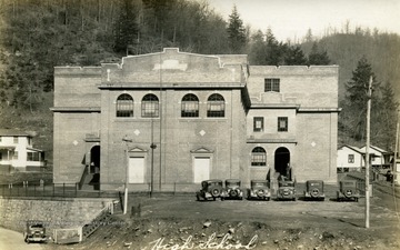 Front view of brick high school with seven, ca. 1930 cars parked outside.