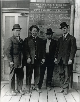 Group portrait of operators in front of a C&amp;O Railway schedule board. L to R; David W. Morgan, Valmer A. Meadow, J. B. Thomas, Todie B. Green. Other information the back of the photograph includes: "from Roy Long Coll 1998"