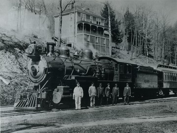Employees stand with a Chesapeake &amp; Ohio (C&amp;O) locomotive sitting on the track in front of the Yellow Goose Boarding House and the McKendree Hospital (a miners' hospital) behind the boarding house. The employees are, left to right: W. L. Burke, Engineer; H. E. McFadden, Fireman;; Jim Johnson, Boy mast(?); Pete Challonen, Dispatcher; D. H. Hontsovln, Conductor; Floyd Lewis, Brakeman.