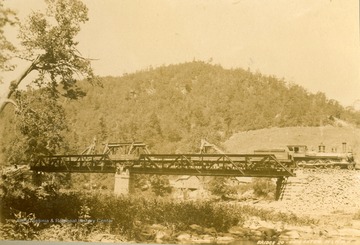 A locomotive, pulling flat cars rolls over  Bridge No. 20. Three unidentified people are standing on the middle pier.