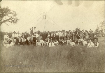 The group of approximately 60 men, wearing bowler, panama and fedora hats, posing with an Irish harp, fiddles, cooking pots and an 8 foot stove-pipe. It was believed to be taken at the Green Sulphur Annual Fair. The man at the extreme right is Timothy Field but all others are unidentified. Other information inscribed on the back includes: "Suspension construction walled circular tent, 2 poles &amp; 4 guide wires, see tent peg at extreme left, Privy at extreme right ...". The photograph was purchased by Stephen Trail in Hinton, West Virginia. 