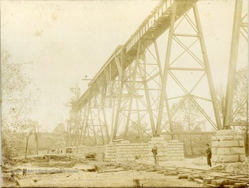 Two unidentified men stand under a partially built trestle bridge during the construction of the Ohio extension of the Norfolk &amp; Western Railroad.