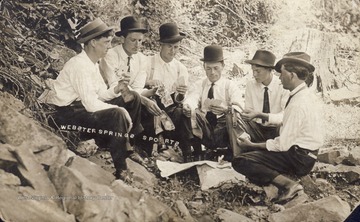 Photograph of poker players outside, with money in hand, huddled around a rock "table." Written on the back of the photograph; "Everete Hawkins of Webster Springs and friends."  Identified individuals are Grover Starcher (second from left) and Elias Hammond (third from left).Courtesy of Steve McElwain, Sutton, Braxton County.