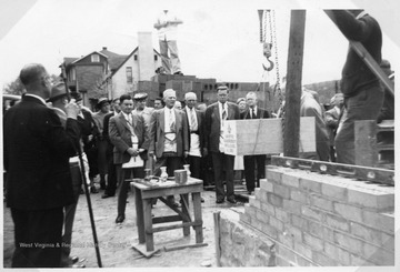 Several unidentified people, some wearing  Masonic aprons, watch as the cornerstone of the new church is put in place during a ceremony. 