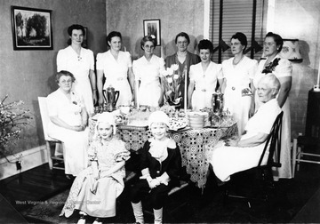 Several ladies gather around a table set for tea and two children sit in front wearing colonial-era costumes. Back row, l to r: Mrs. Baker; unknown; unknown; Mrs. Carl Walker; and Mrs. Welch. End of table, l: Mrs. Helen Camp; other end, Mrs. W. B. Bartlett. In front on left: Alece Fisher.