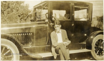 George Barrick Sr. seated on the running board of a automobile. The passenger in the vehicle is unidentified. 