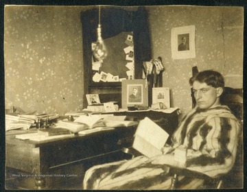 Unidentified male student with a dazed look and wearing a robe, holds an open book at a desk. 