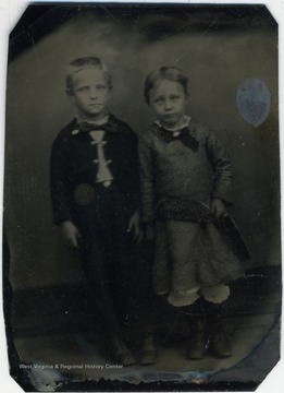 Young girl and boy (around five or six years old). The boy is in a suit and bow tie while the girl is wearing a knee length dress with laced trimmed bloomers. 