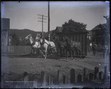 A team of four horses, hitched to a small building, pull it through town streets. Subjects are not identified.