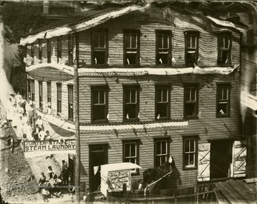 Several people, including workers and children line the sidewalk on the left side of the building which is decorated with bunting and flags. A delivery wagon, hitched-up and covered with advertisements is parked in front.