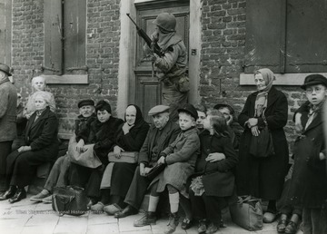 Information on back of photo reads: "German civilians sit with their children outside of a house in a Reich town captured by troops of the Ninth U.S. Army advancing to the Rhine River. The civilians have been lined up for questioning by an American officer. Units of the Ninth Army reached the Rhine March 2, 1945, when they captured Neuss opposite the industrial center of Dusseldorf."