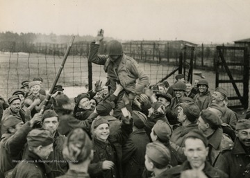 Information on back of photo reads: "Liberated Russians cluster around a Ninth U.S. Army soldier, carried high on their shoulders, for cigarettes, which they had not seen in many months. When the American finally convinced the Russians that he had no more, they "chaired" him and carried him around the yard before their former prison, the Nazi Stalag 326, south of Bielefeld. The first U.S. troops reached Stalag 326 April 2, 1945. Nine thousand Russian prisoners of war were liberated but thousands were at the point of starvation. Tubercular patients numbered 1,350. in vast mounds all around the camp, 30,000 Russians, most of them starved to death, were buried in heaps of 500. Major Gregory Matviev, who was captured in Sebastopol in 1942, reported that hundreds died daily of starvation and "about 50 were shot every other day for no reason at all.""