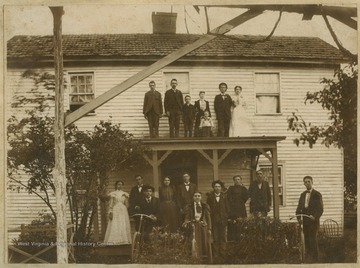 A family poses in front of a house, some are perched on the porch roof.  Three in the front row hold the handle bars of bicycles. Only identified subjects are Samuel A. and Margaret Menear Dill, standing far right under the porch roof.