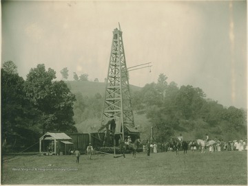 Several people gather to observe the workings of an oil pumping derrick. Note the dog mounted on the horse, center-right.