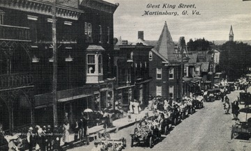 Parade going down the road with U.S. flags attached to the cars driving one after another. (From postcard collection legacy system.)