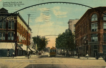 Trolley travels down middle of road underneath lighted archways as people go about their day. See original for correspondence. (From postcard collection legacy system.)