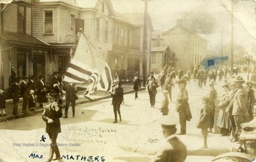 Max Mathers leads the procession carrying the American Flag. See original for correspondence. (From postcard collection legacy system.)