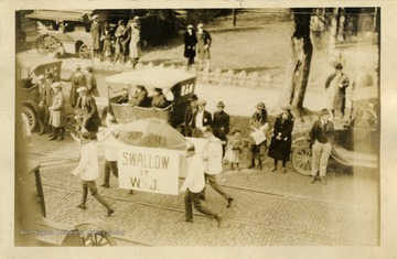 Several students carry a fake pill with a sign that reads "Swallow it W.J.". (From postcard collection legacy system.)