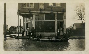 People stand outside of an office building while others sit in a boat on the flooded street.(From postcard collection legacy system.)