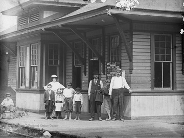 Group of people stand outside of railroad depot. Departure times for several trains on the wall behind men on the right.