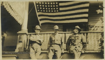 Three uniformed men stand below a large American flag hung from a home's porch. 