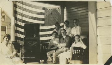 A group is shielded from the sun by a large American flag as they enjoy the porch swing and music.