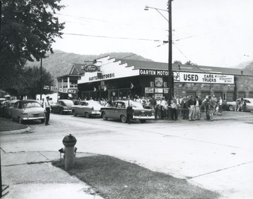 A crowd gathers around the new Ford automobiles parked in front of the store lot.